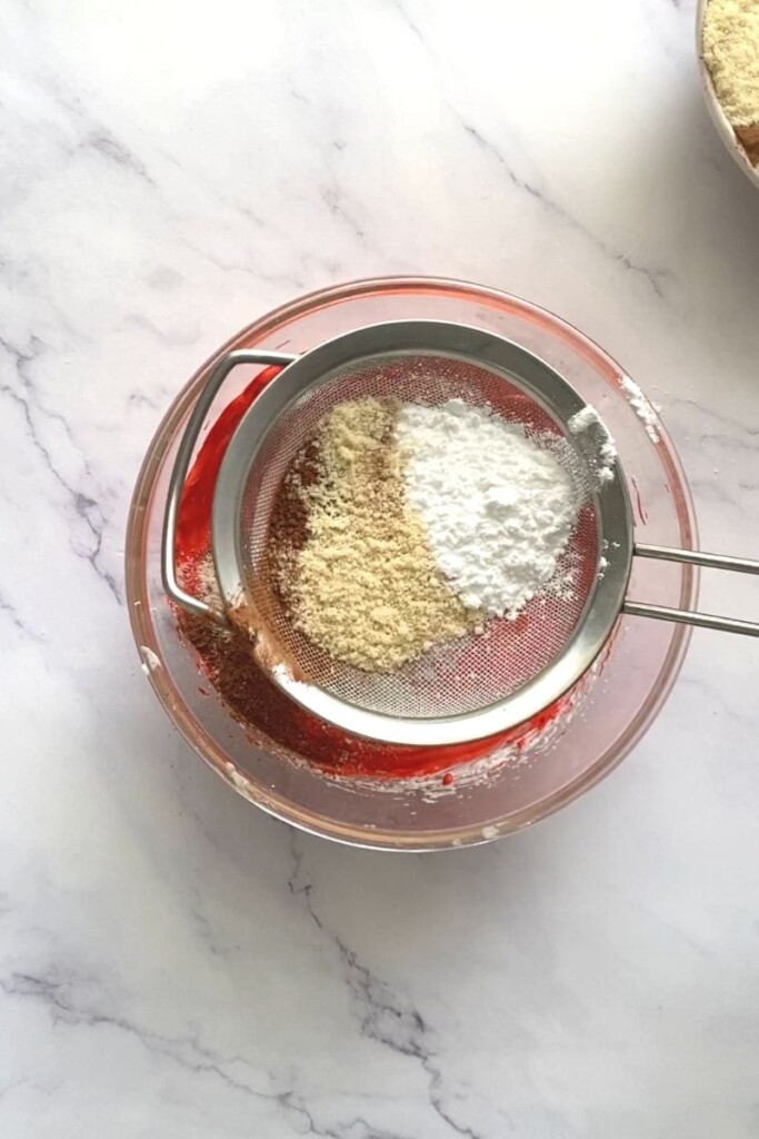 Almond meal, powdered sugar and cocoa powder in a sieve on top of a bowl of red meringue.