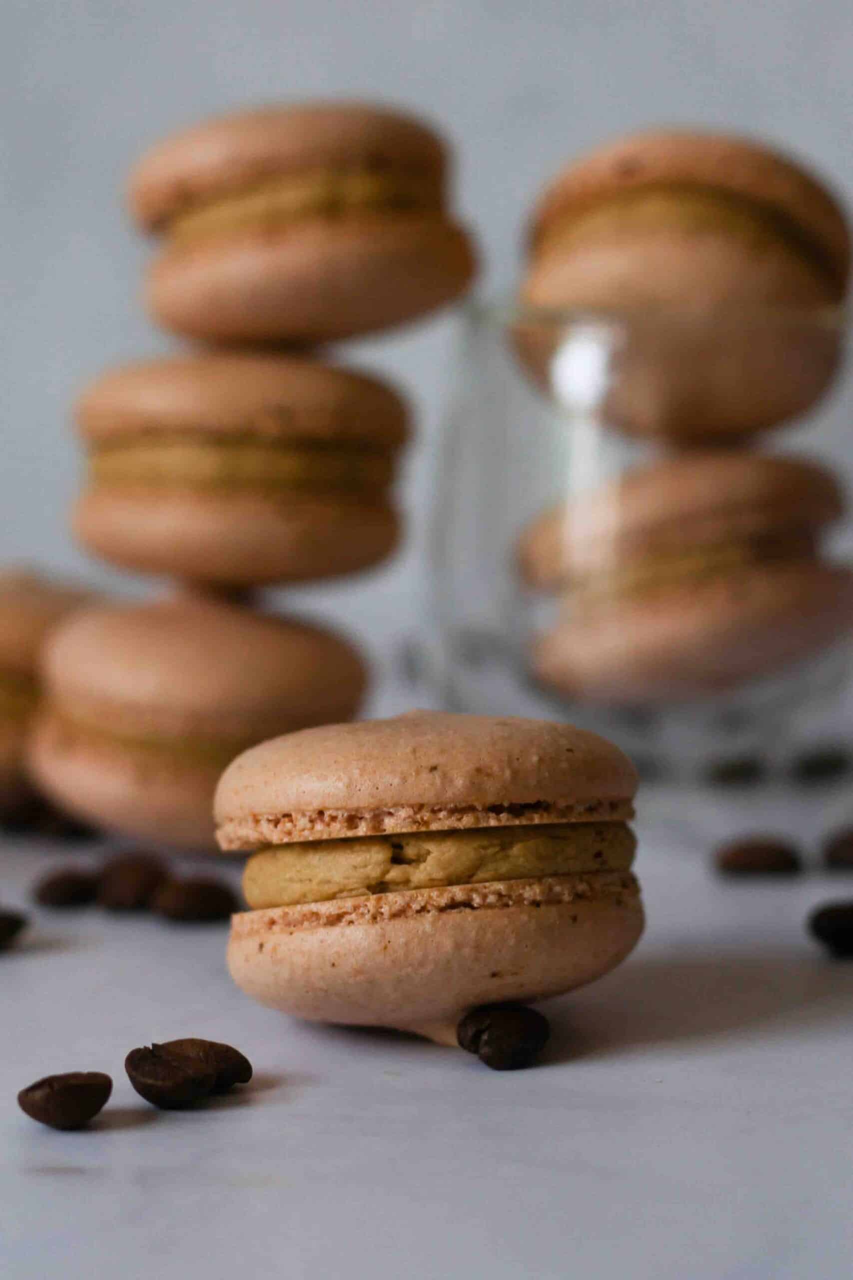 Coffee macarons with macarons stacked in the background and a close up off one in the front.