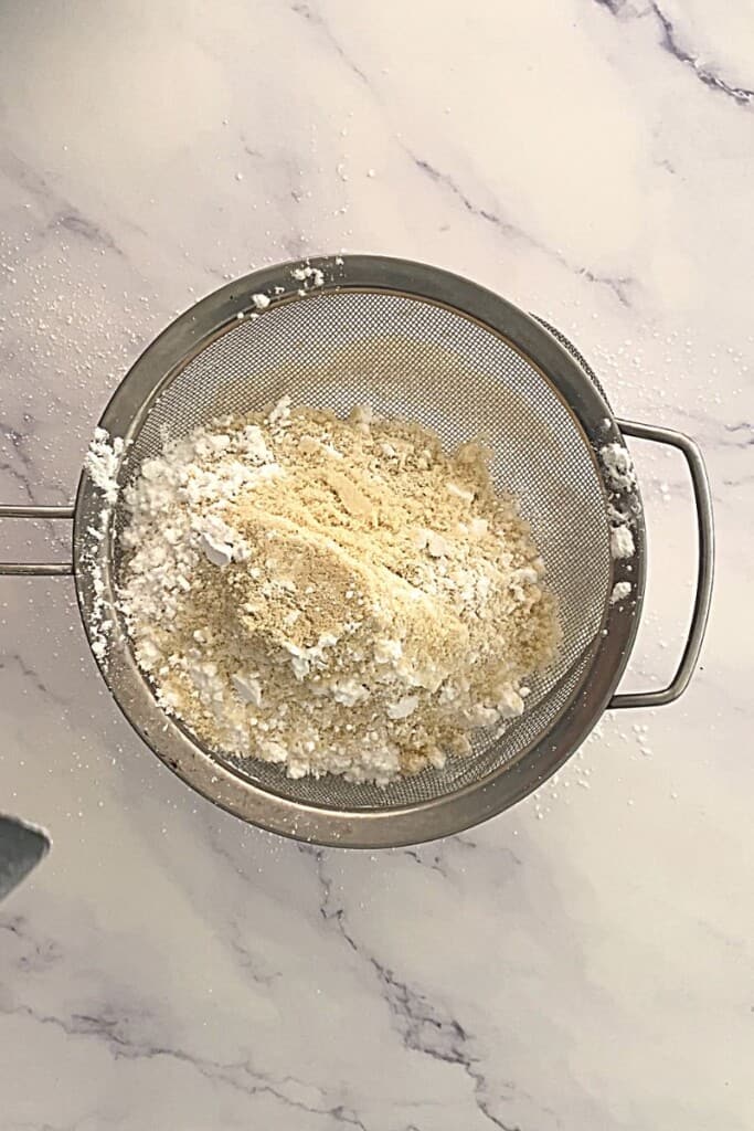 Almond meal and powdered sugar being sieved on meringue.